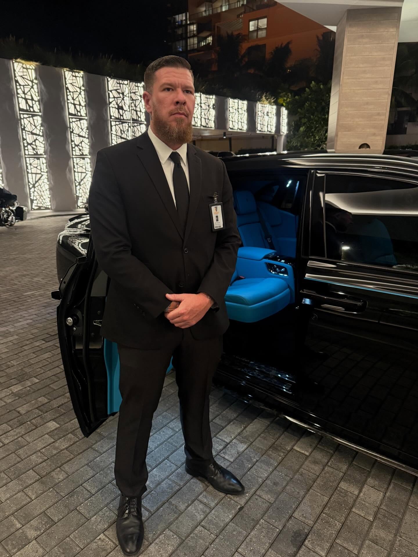 Suited Spectrum agent standing post at the open door of a black Rolls-Royce at a Miami residence